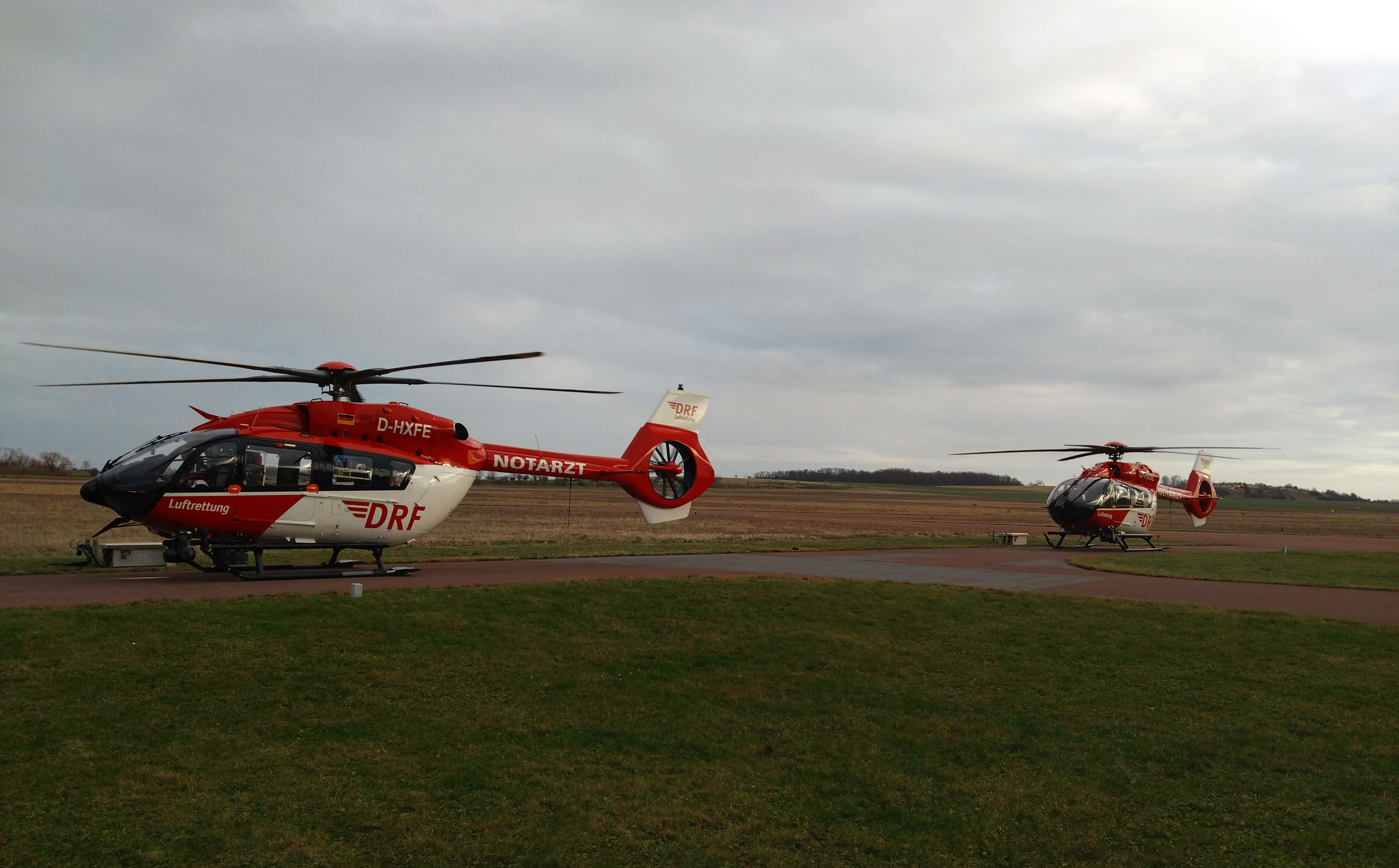 Zwei rot-weiße Rettungshubschrauber der DRF Luftrettung stehen abflugbereit auf einem Vorplatz eines Flugplatzes.