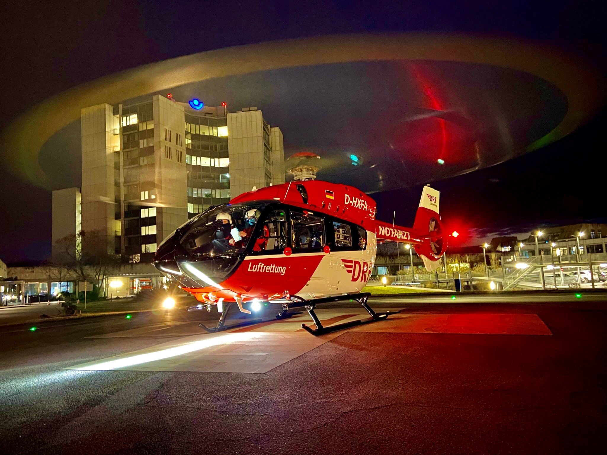 Ein rot-weißer Hubschrauber steht mit drehendem Rotor auf einem Kliniklandeplatz in der Nacht.