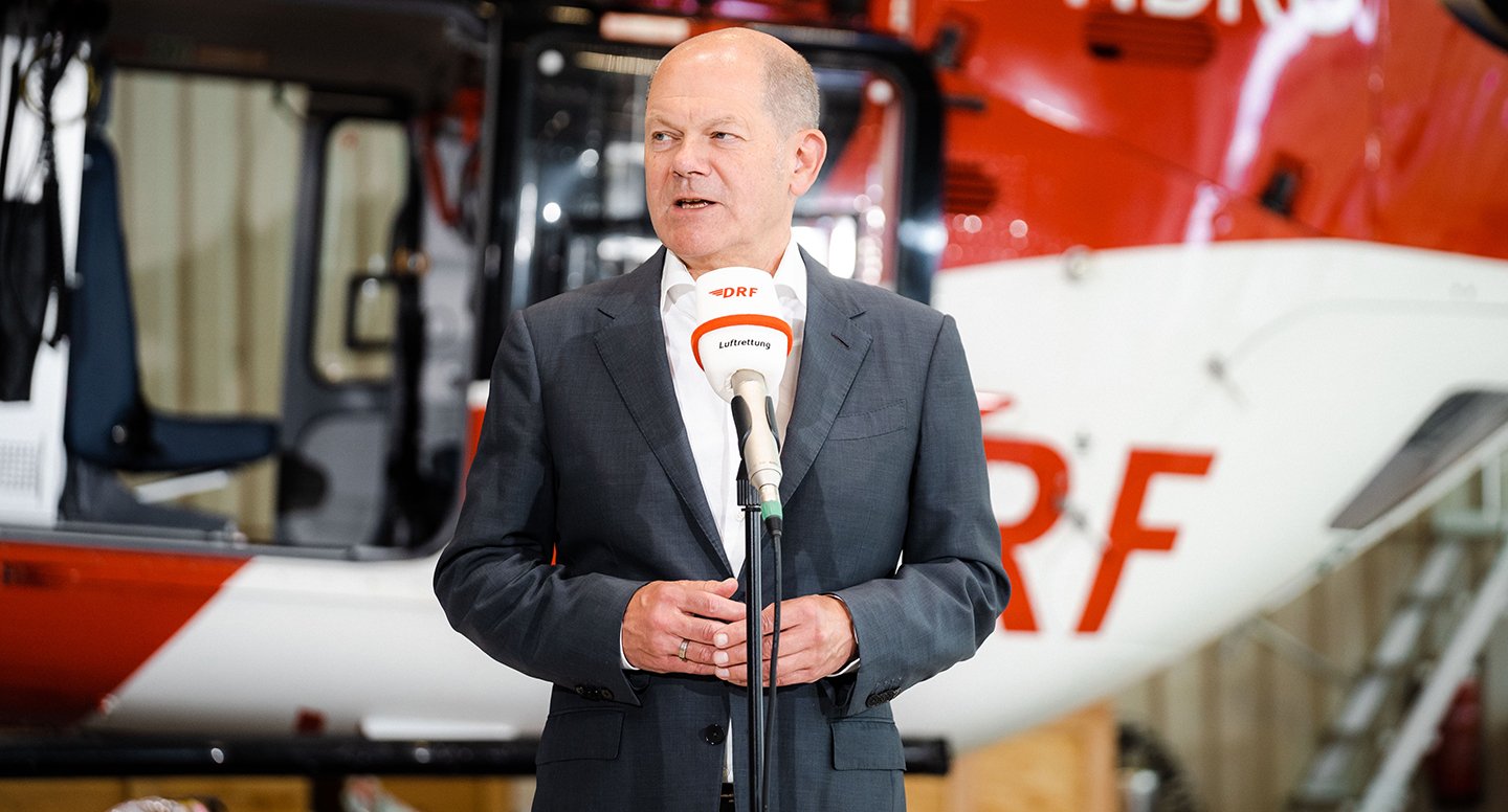 Bundeskanzler Olaf Scholz steht vor einem Mikro und spricht. Im Hintergrund steht ein Hubschrauber.