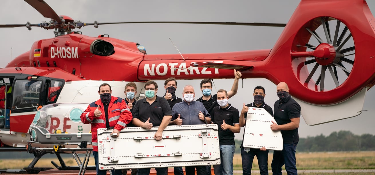 Mehrere Personen stehen vor einem rot-weißen Hubschrauber und halten eine Bodenplatte für das EpiShuttle vor sich.