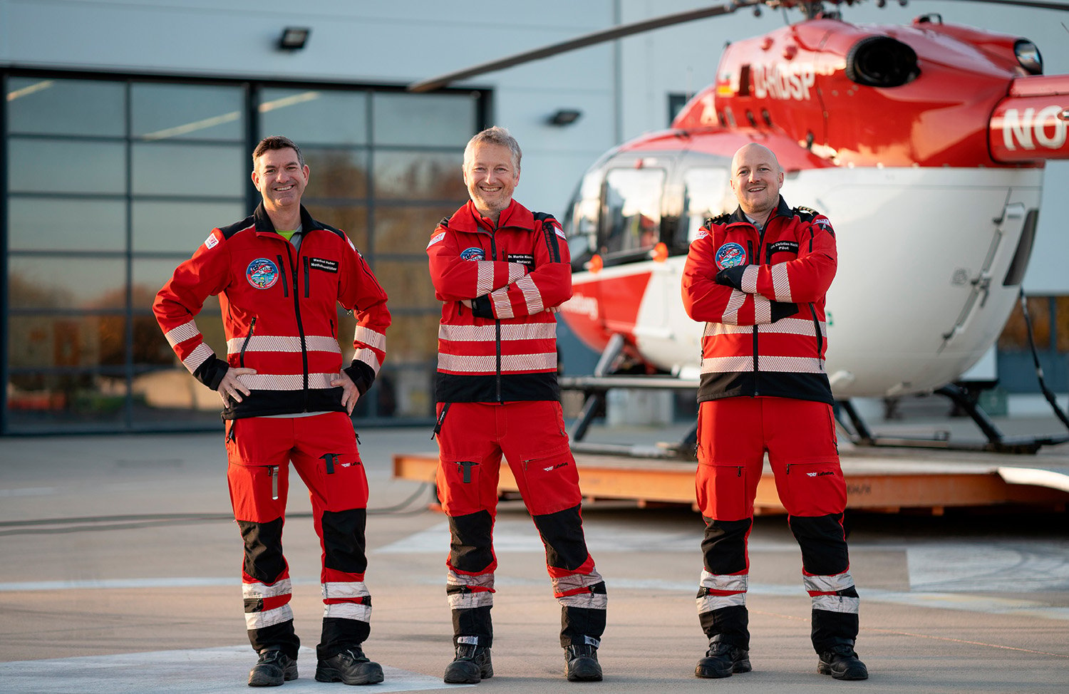 Drei Männer in Rettungsuniform stehen nebeneinander vor einem Hubschrauber